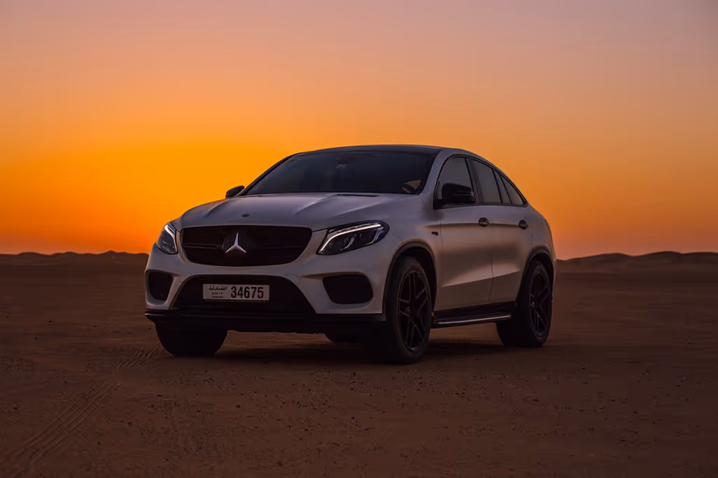 White Mercedes-Benz SUV parked on desert terrain at sunset with an orange sky.