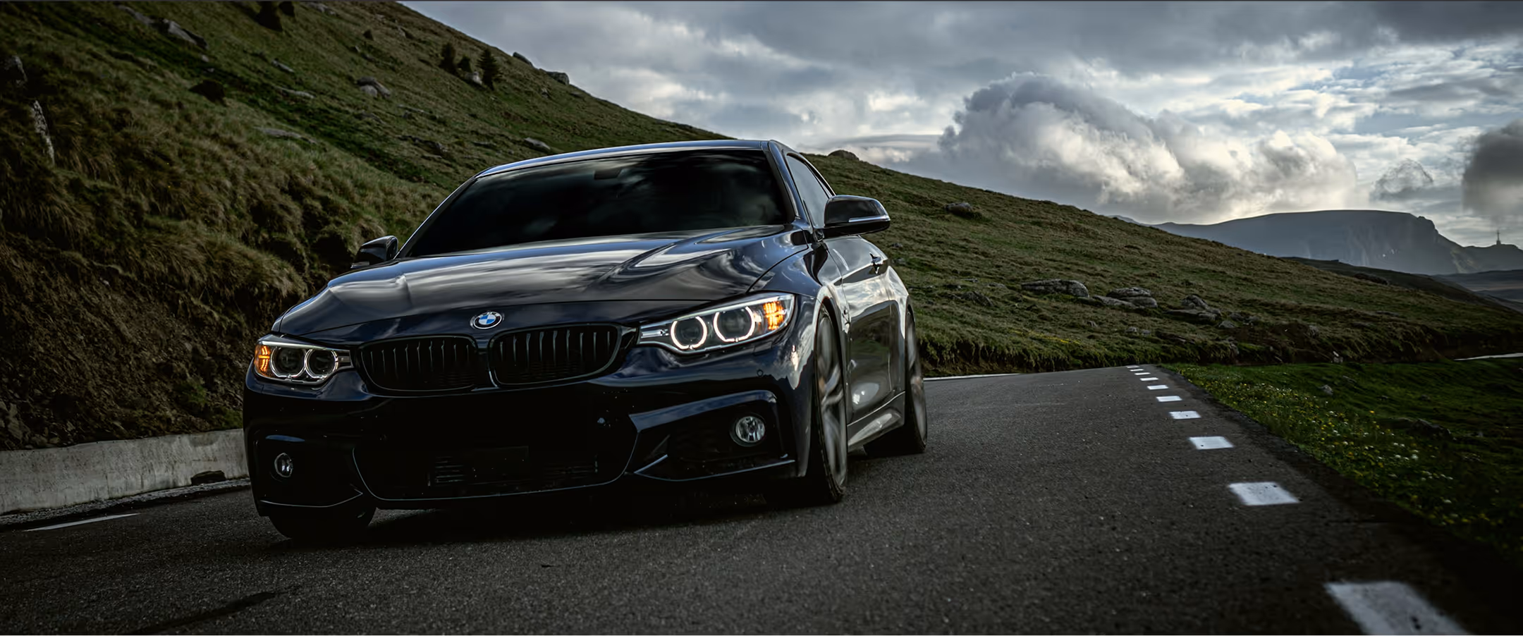 Black BMW car driving on a winding mountain road with grassy hills and cloudy sky in the background.