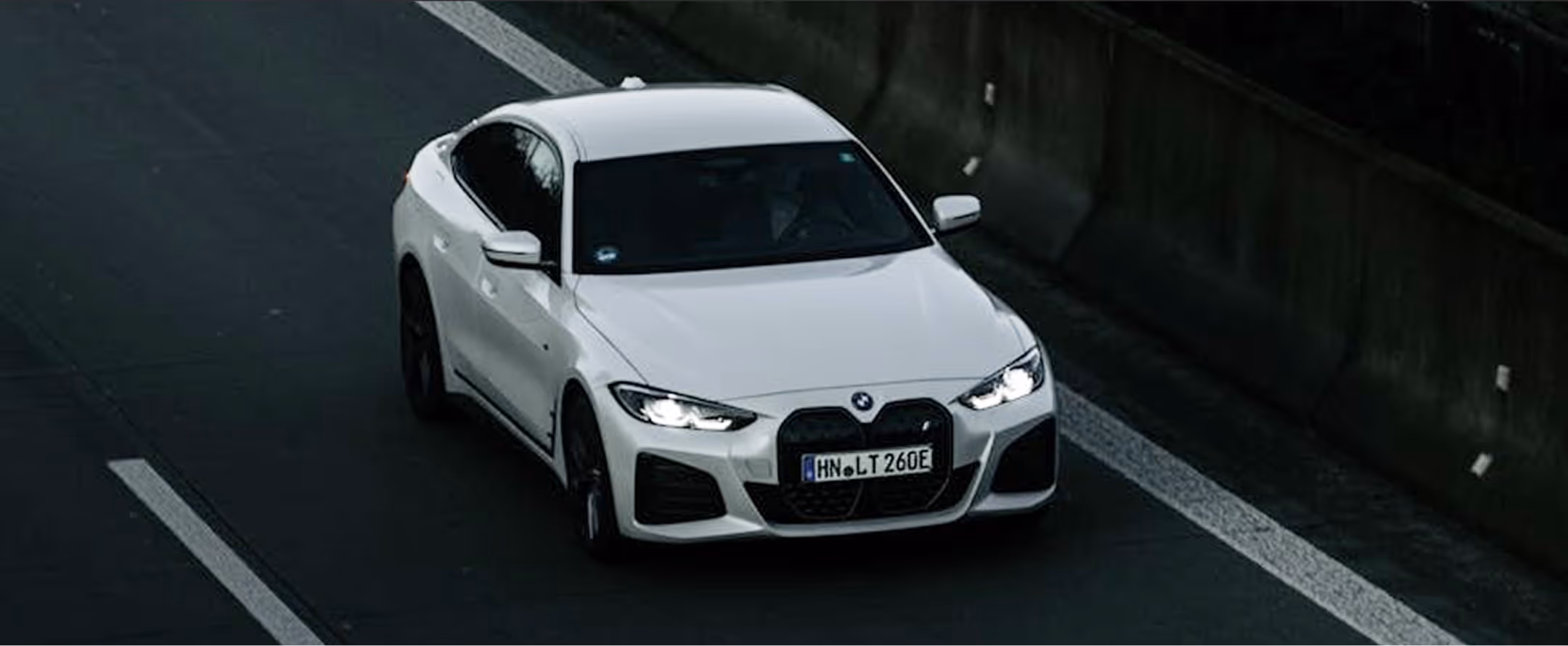 White BMW car driving on a dark road with headlights on next to a concrete barrier.