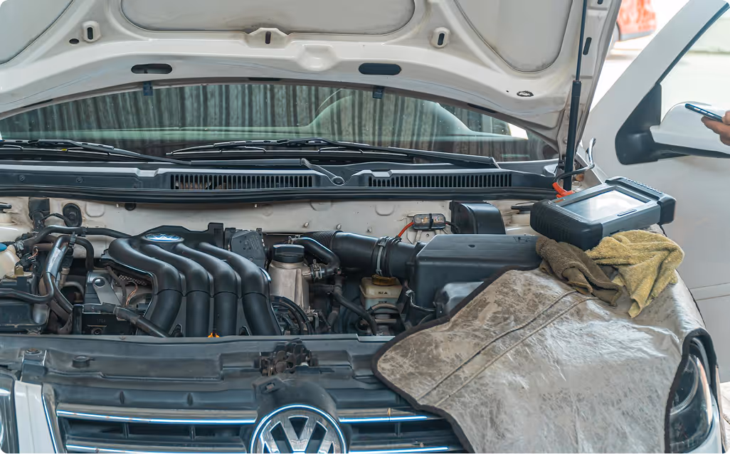 Open hood of a Volkswagen vehicle showing the engine with a diagnostic device and cloths on a mat.