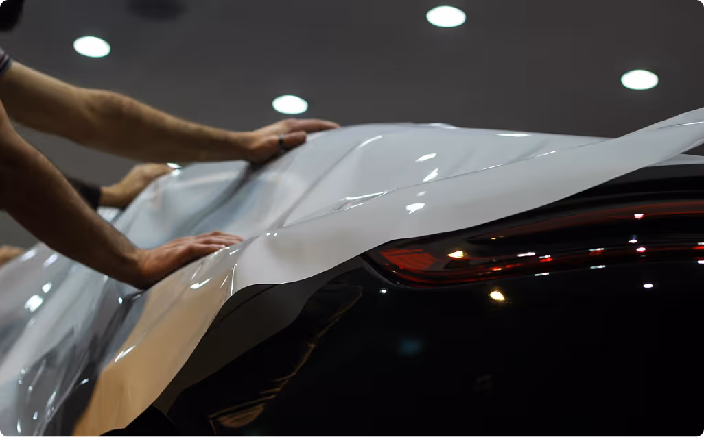 Hands applying a white vinyl wrap to a black car under bright ceiling lights.