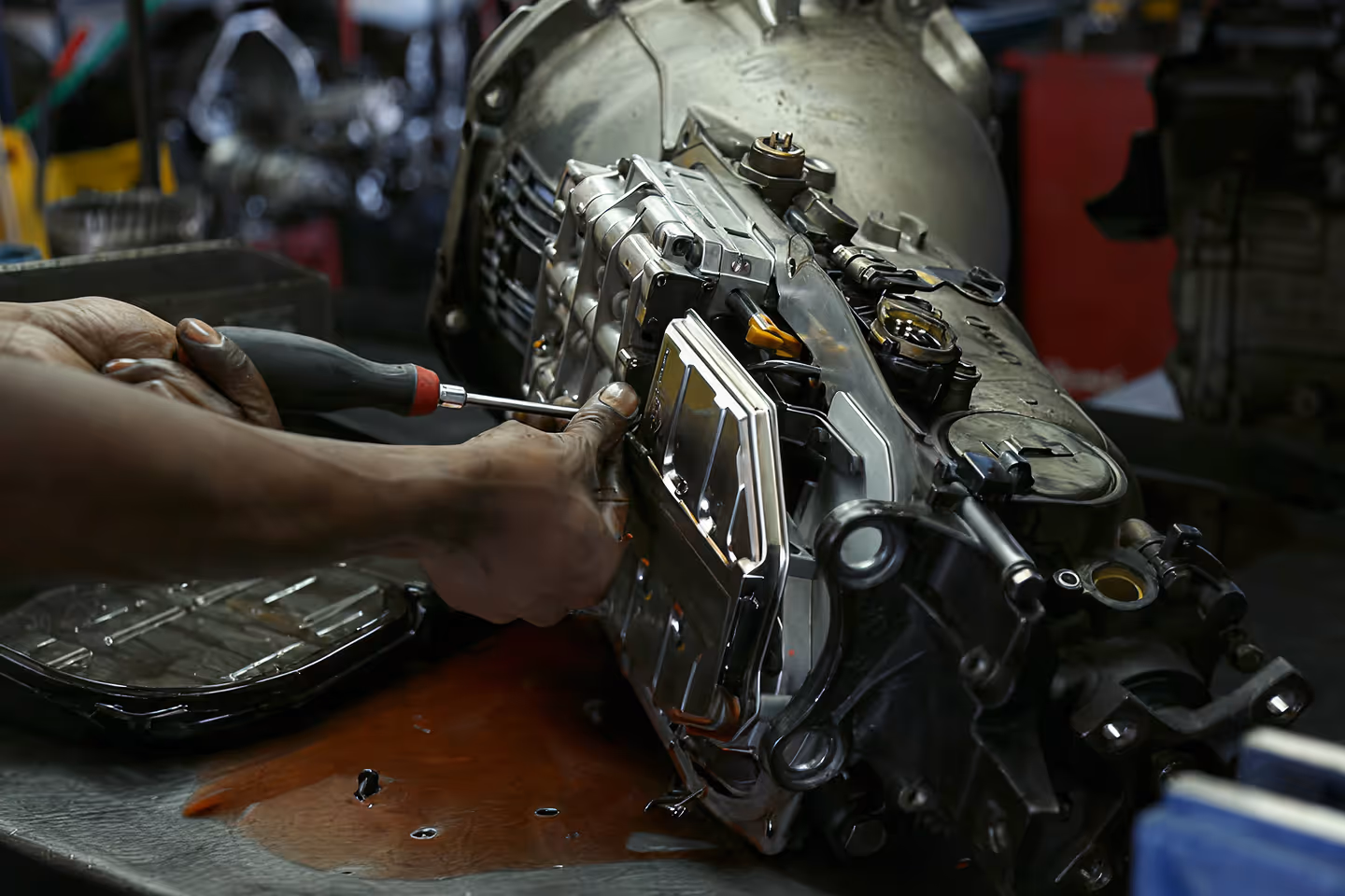 Mechanic's hands using a screwdriver to repair a large, disassembled automotive transmission on a workbench with fluid spills.