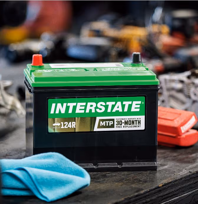 Interstate car battery with green top and label showing 30-month free replacement warranty on a workbench with tools and a blue cloth nearby.