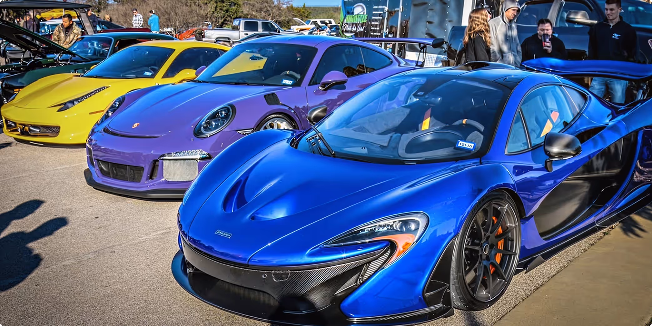 Three sports cars parked side by side at a car show with people in the background, featuring a blue McLaren in the foreground, a purple Porsche, and a yellow Ferrari.