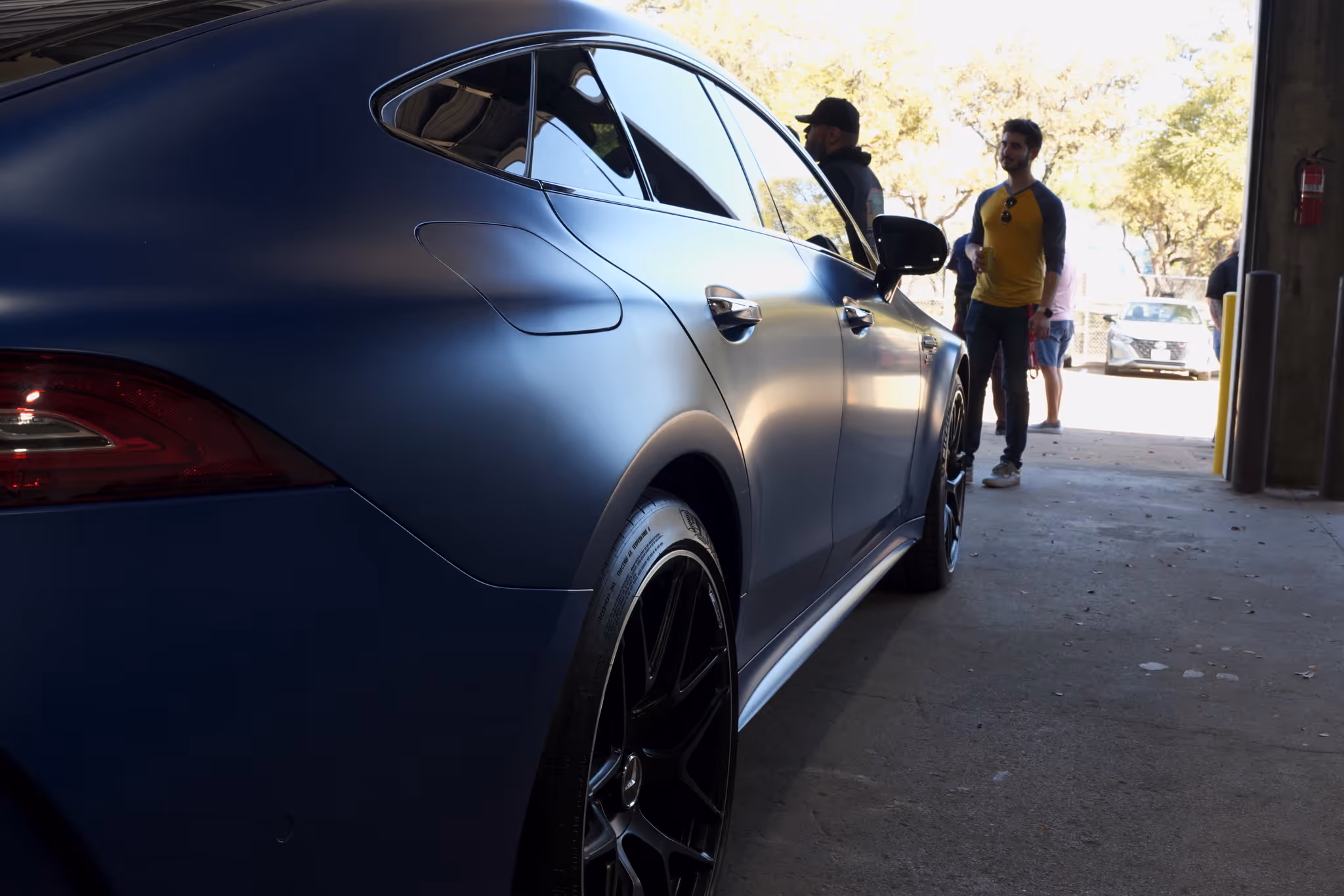 Side view of a matte blue luxury car parked inside a garage with people standing and talking in the background.