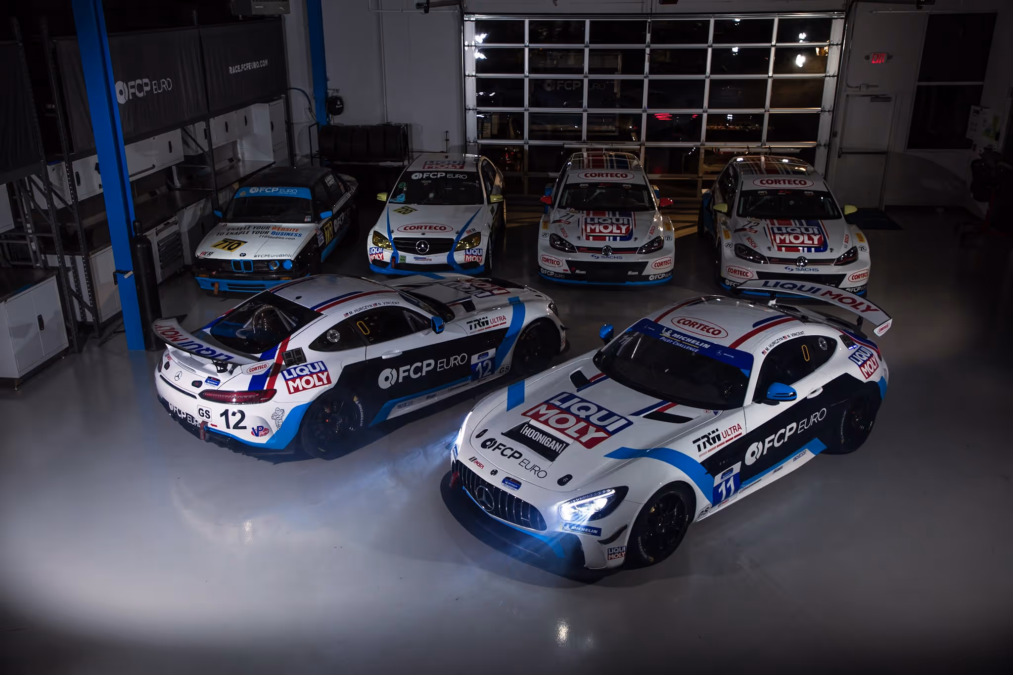 Six white and blue FCP Euro race cars parked inside a garage with logos on body panels.