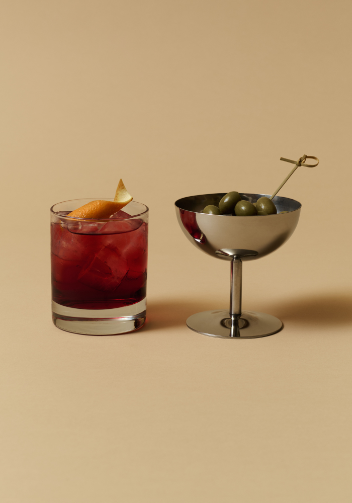 Un cocktail Negroni rouge rubis et une coupe remplie d'olives sur un pic, présentés sur fond beige. Art de vivre avec Anchor.