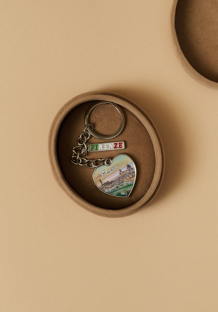 Coffret rond en cuir ouvert contenant un bijou de sac en forme de cœur argenté sur fond beige. Détail d'artisanat avec Anchor.