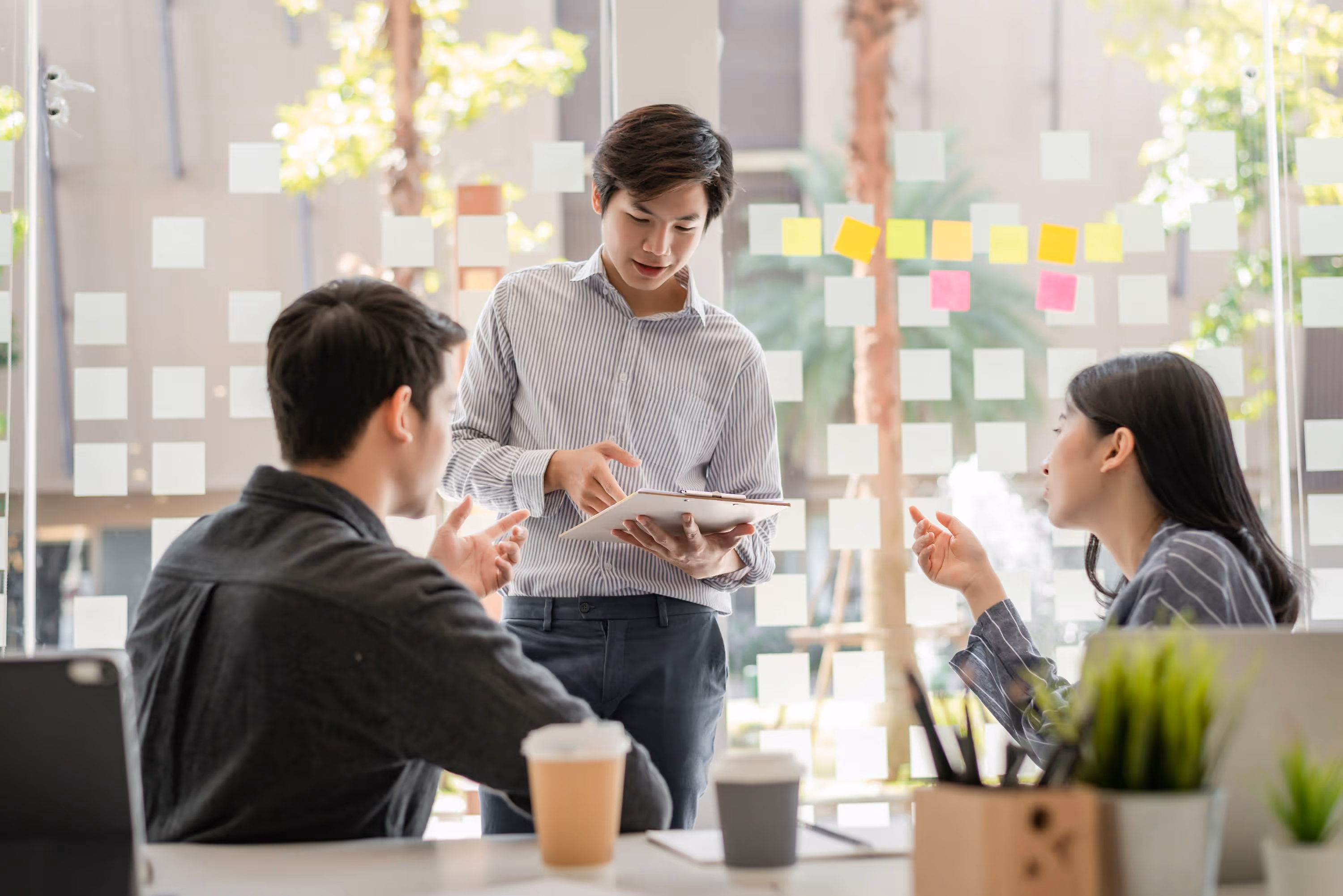 Three colleagues discussing work in an office with sticky notes on the glass wall behind them.