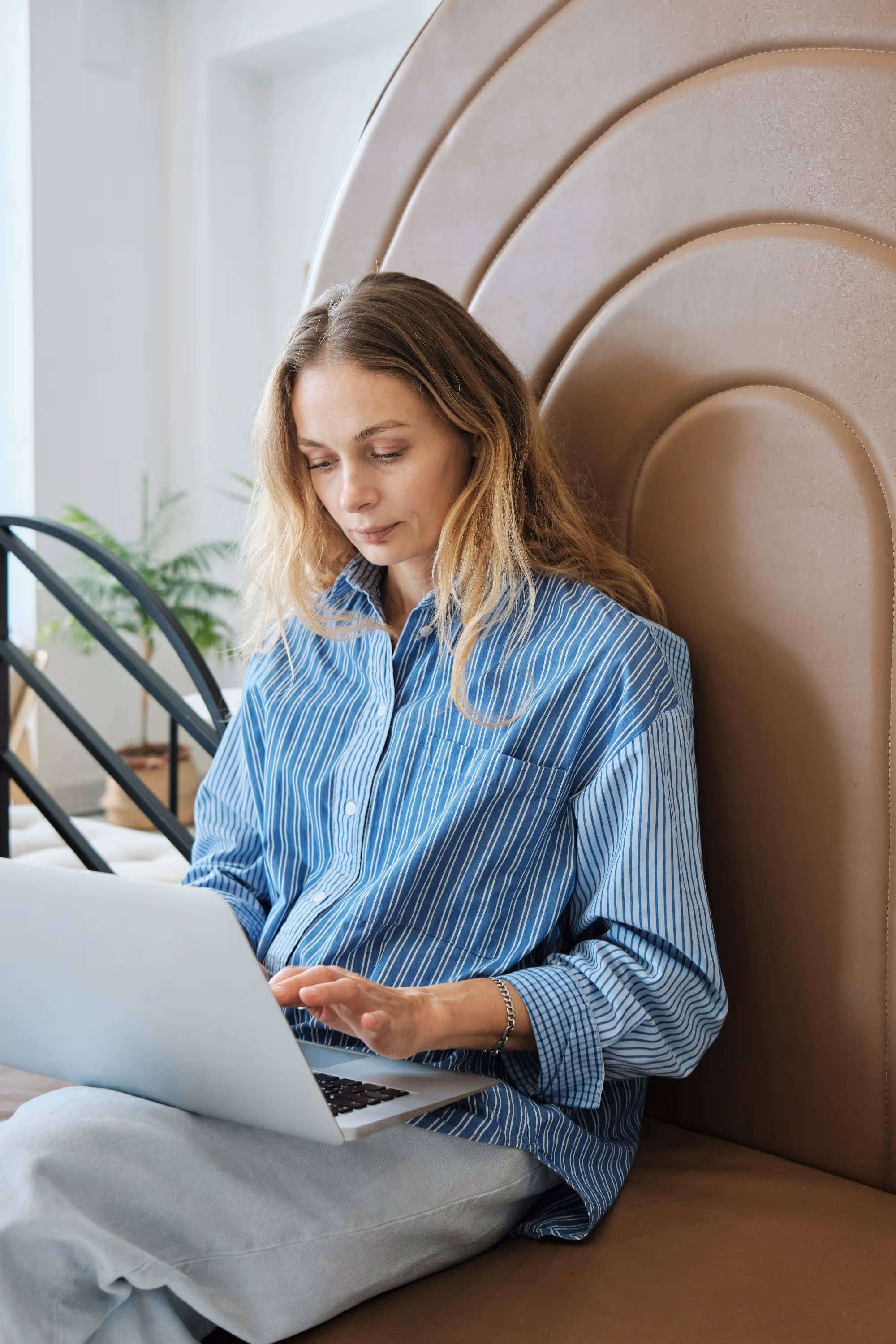 Woman in blue striped shirt sitting on a brown cushioned bench using a laptop.