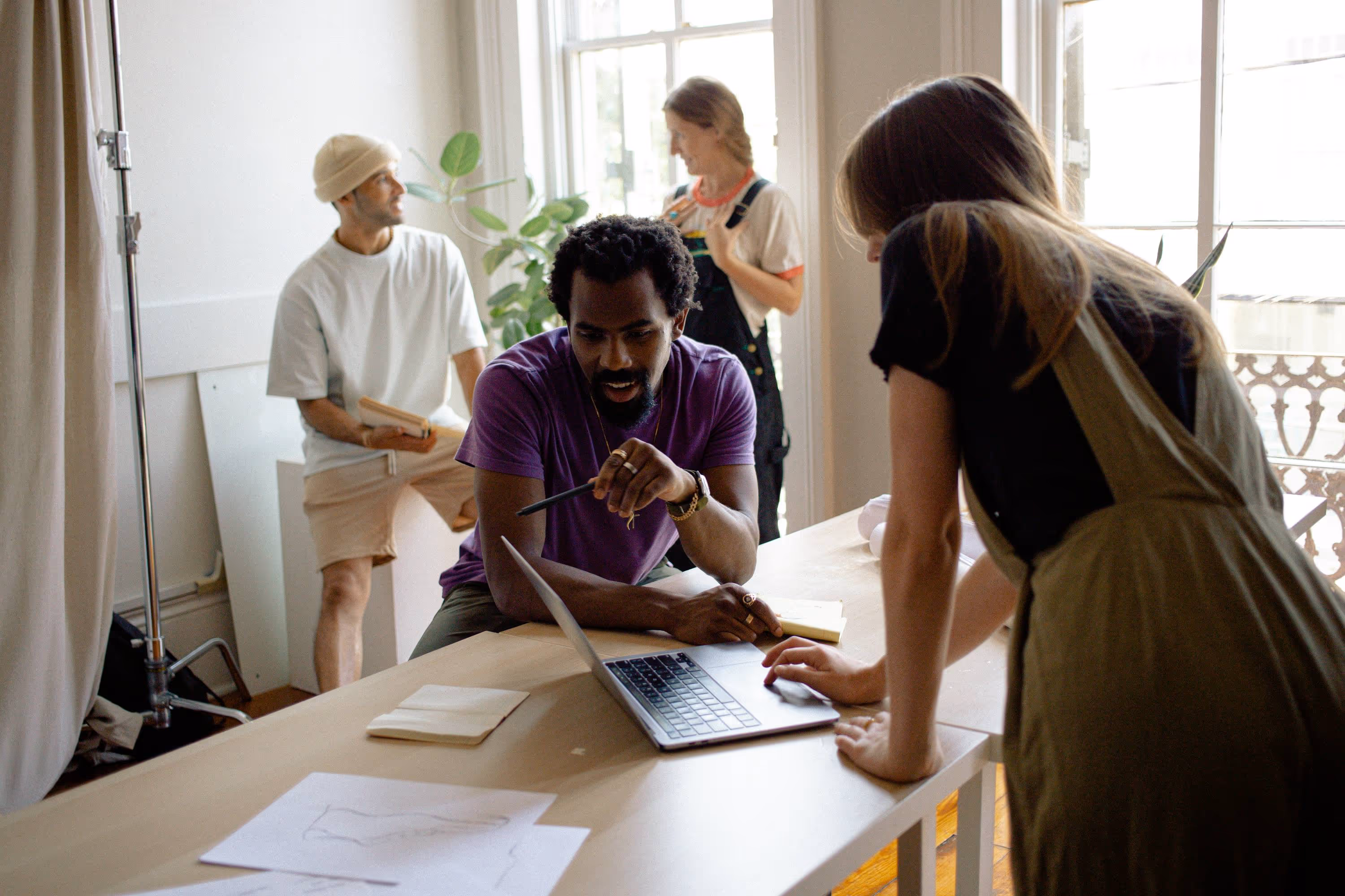 Four people working together in a bright room, two looking at a laptop on a table while two others stand and converse near a window.