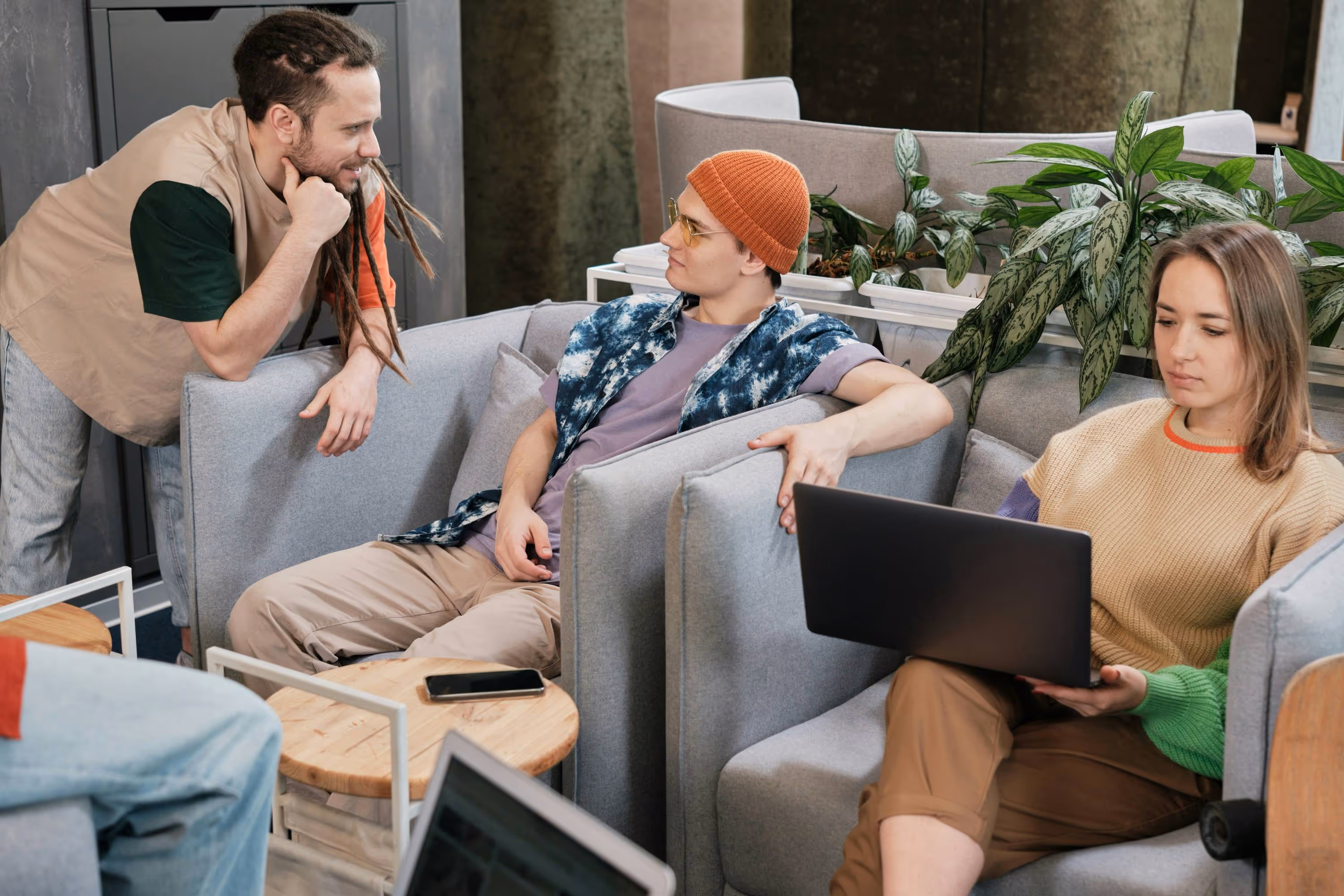 Three young adults in casual clothes sitting and talking in a modern lounge with laptops and a phone on a table.
