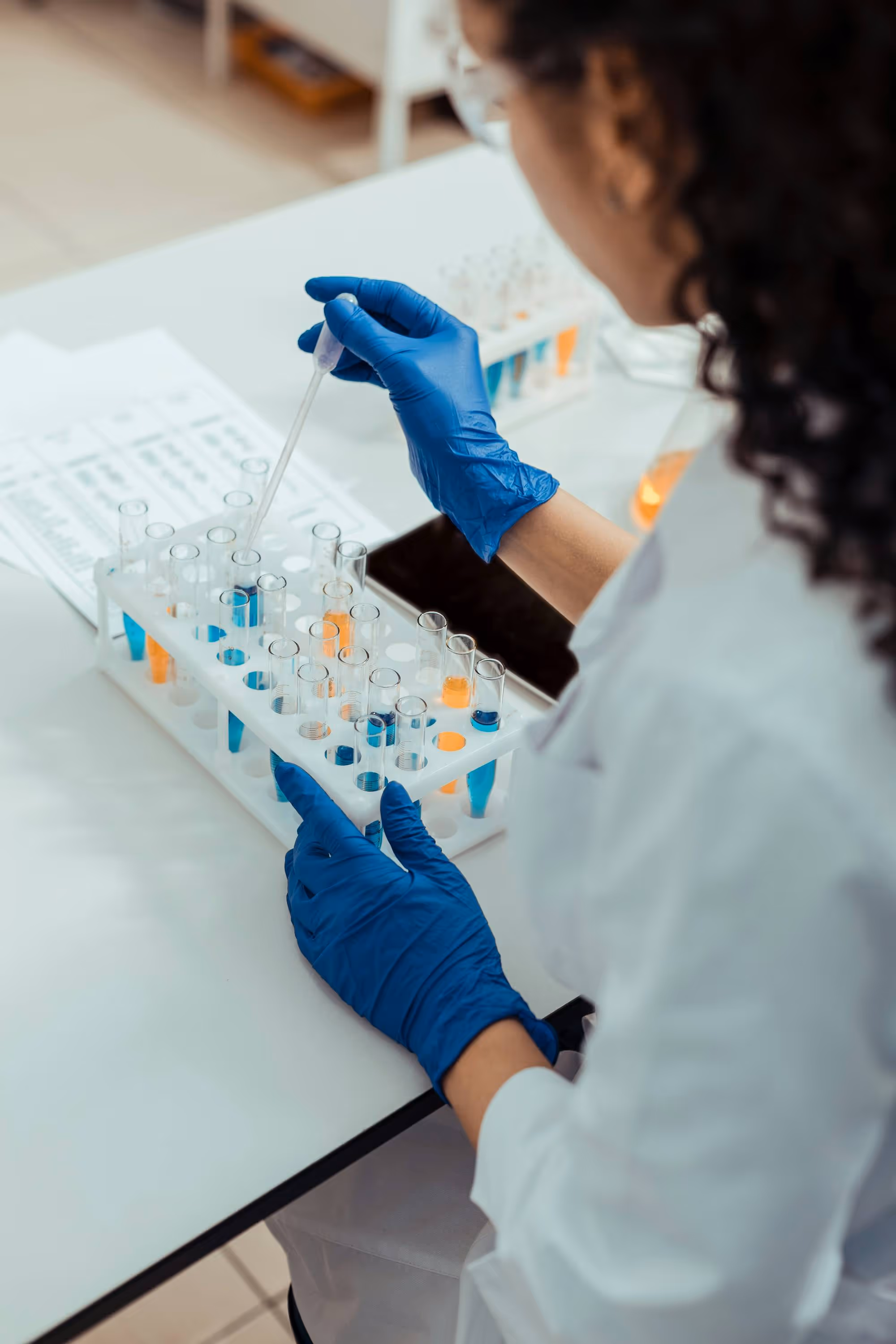 Scientist in a lab coat and blue gloves handling test tubes with colorful liquids using a dropper.