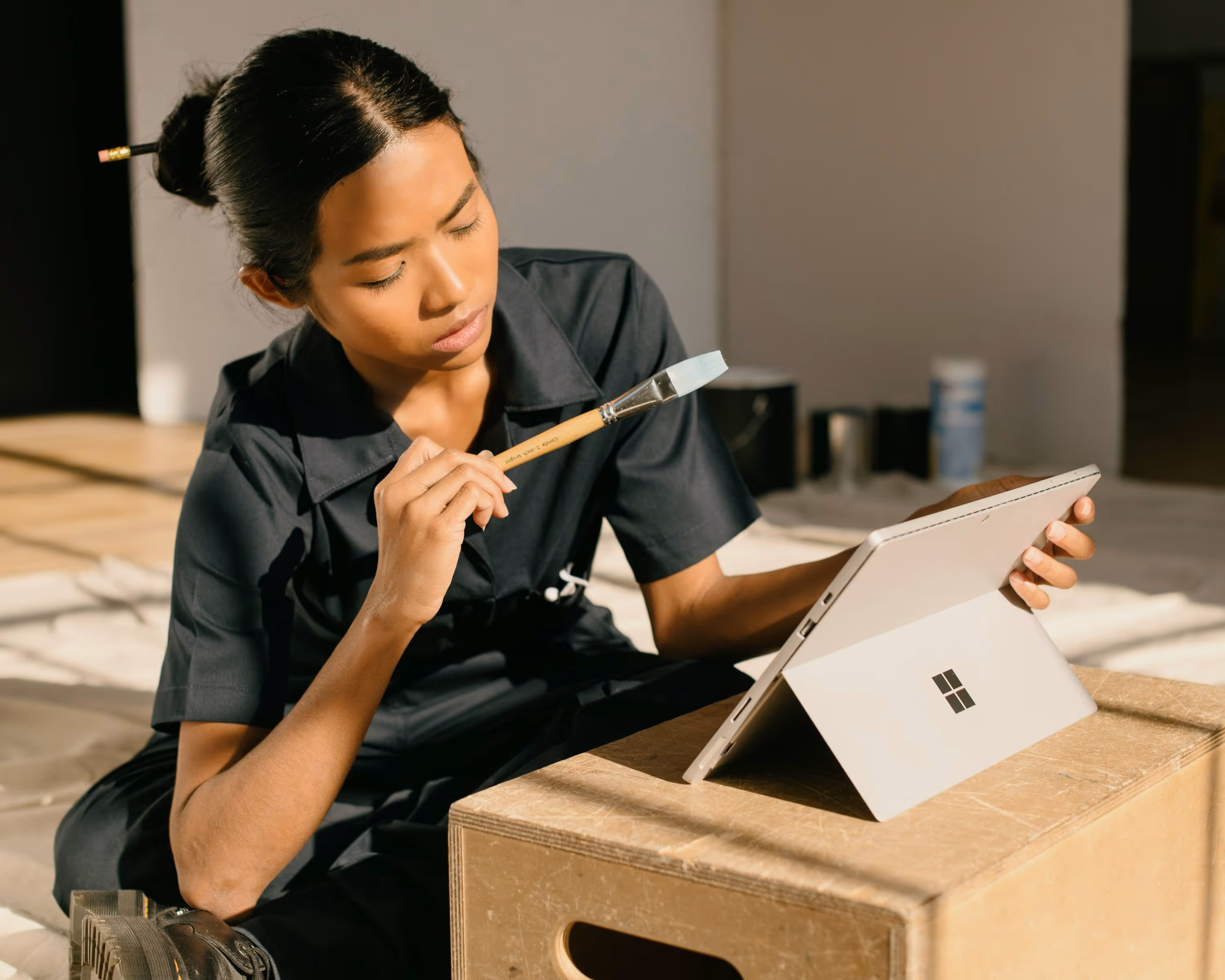 Woman holding a paintbrush and looking thoughtfully at a tablet on a wooden box in a sunlit room.