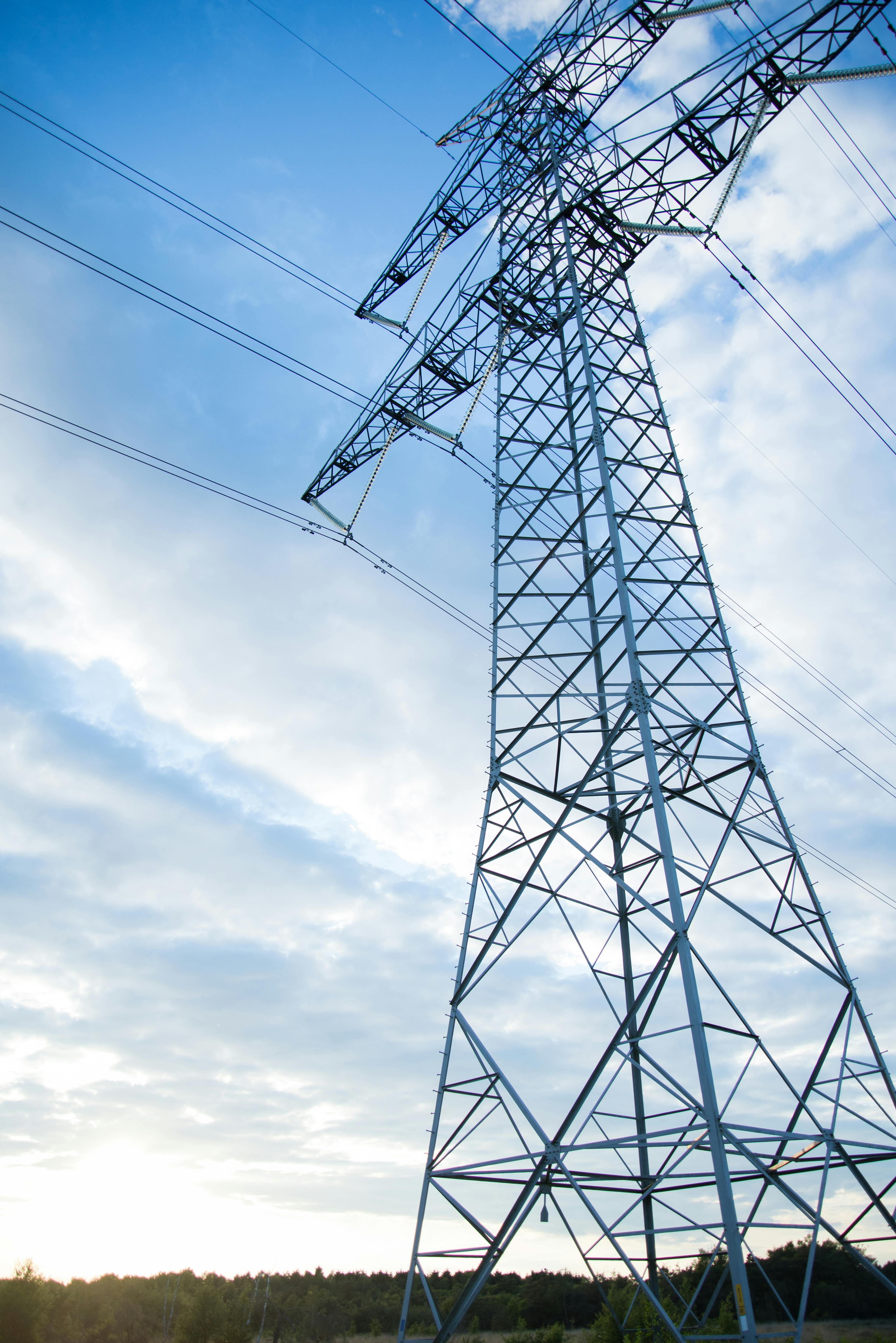 Tall electrical transmission tower against a partly cloudy blue sky over a forested landscape.