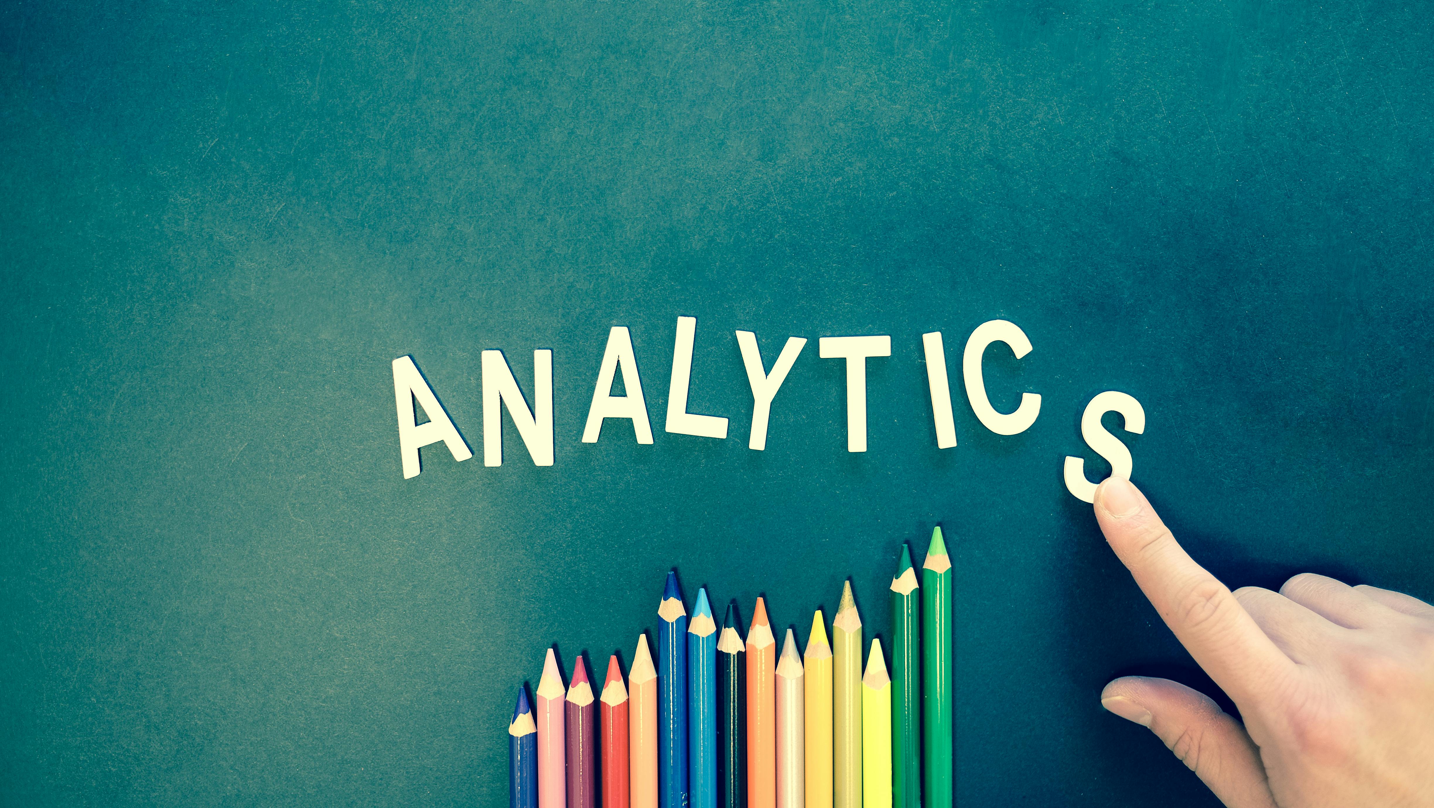 Hand placing the letter S to complete the word ANALYTICS above a row of colored pencils arranged by height on a teal background.