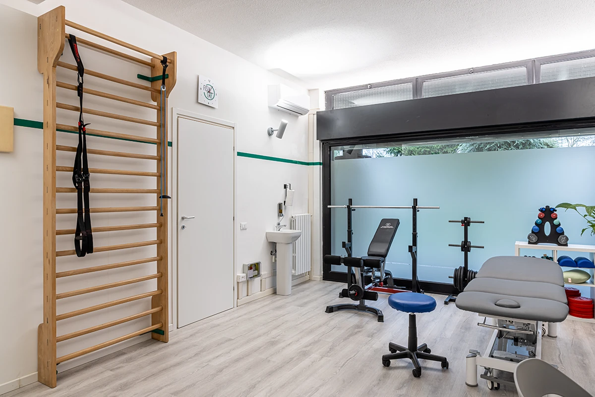 Studio con attrezzature per fisioterapia tra cui una parete attrezzata in legno, panca per esercizi con bilanciere, sgabello blu e lettino da trattamento grigio.