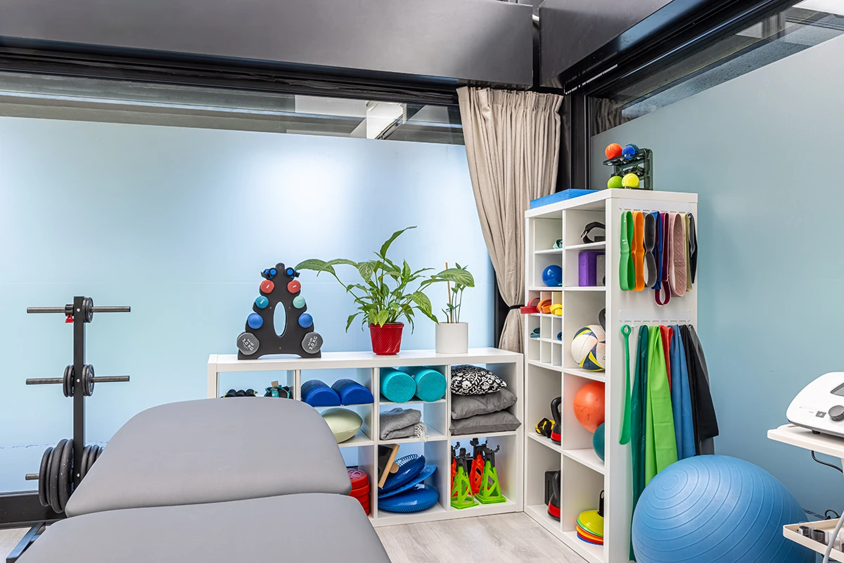 Stanza di fisioterapia moderna con lettino, pesi, elastici, palloni e attrezzi per esercizi organizzati su scaffali bianchi.