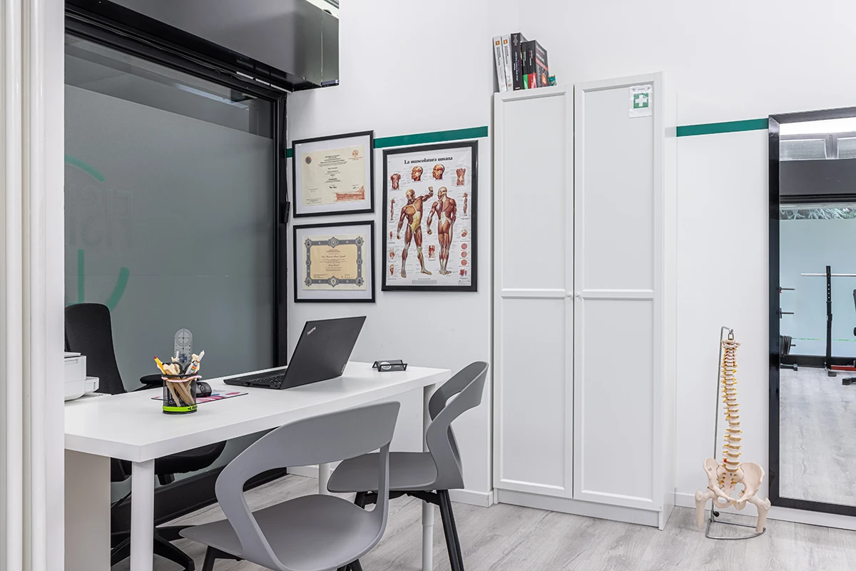 Ufficio medico con scrivania bianca, due sedie grigie, laptop aperto, modelli anatomici, armadio bianco, poster muscolare e scheletro umano in plastica.