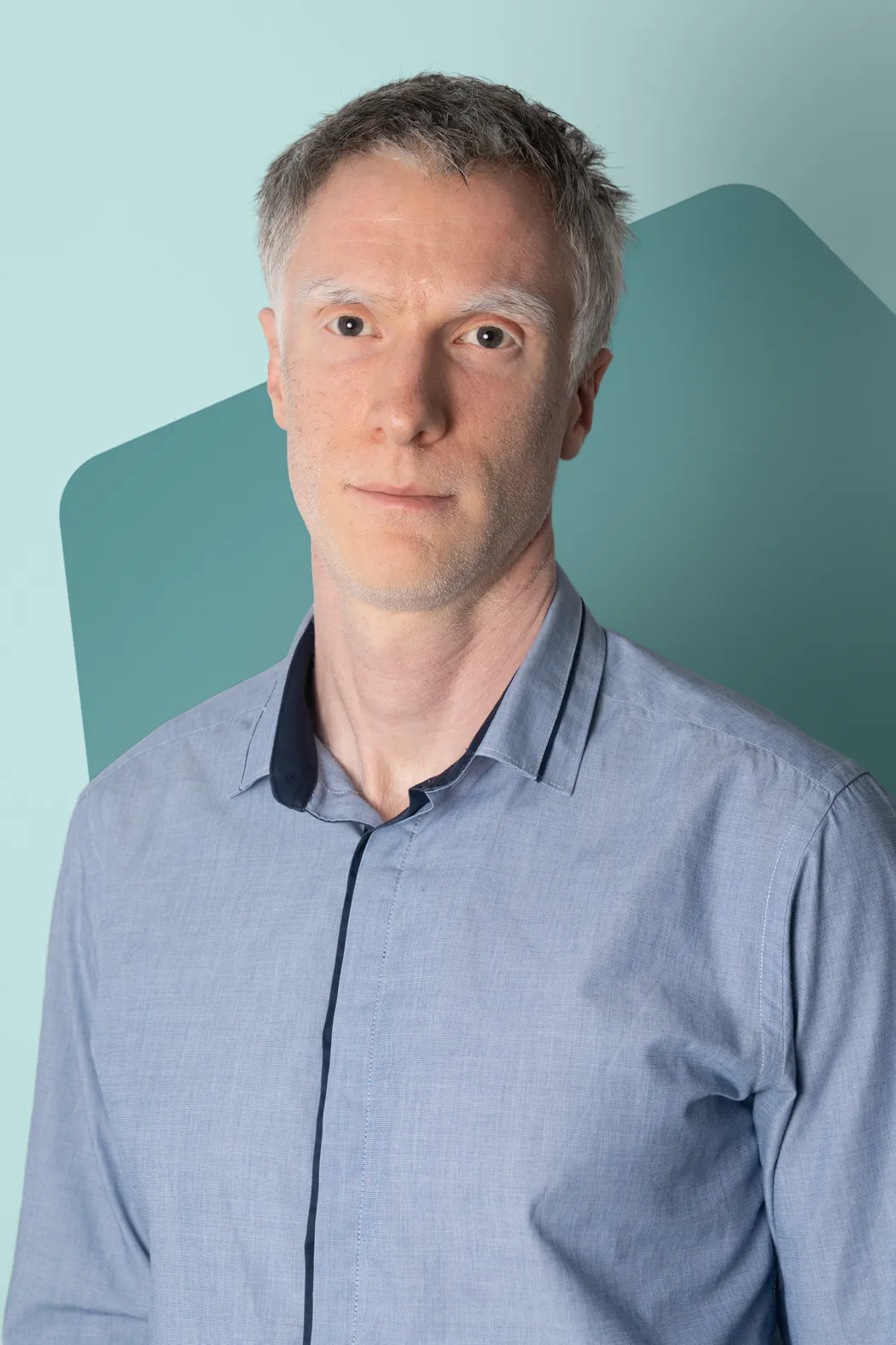 Man with short gray hair wearing a light blue button-up shirt, standing in front of a geometric teal and light blue background.