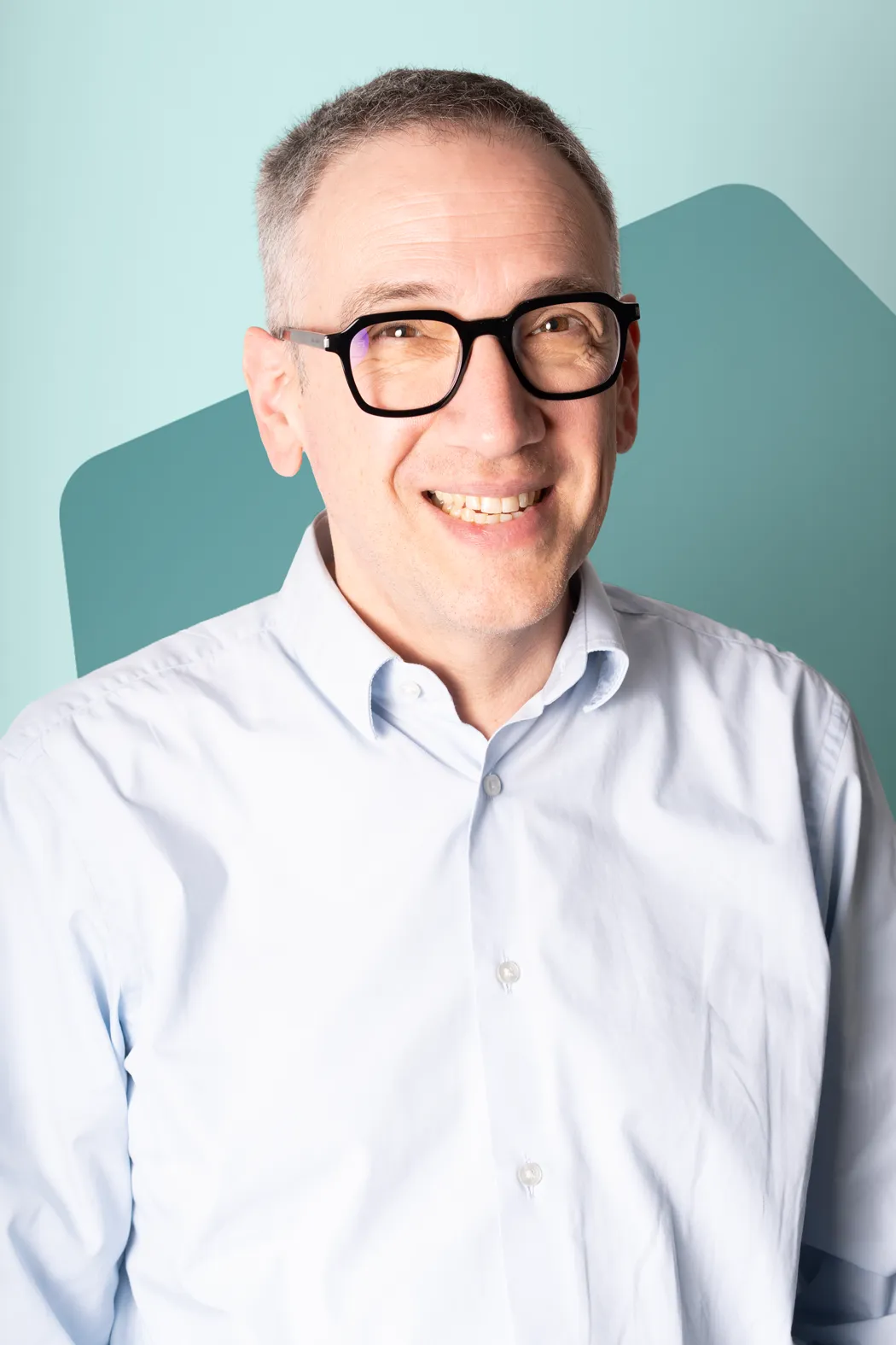 Smiling middle-aged man with short gray hair and black glasses wearing a light blue button-up shirt.