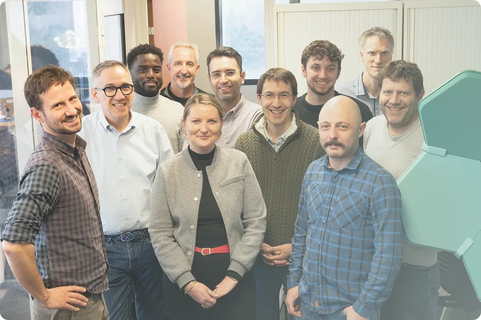 Group of eleven adults smiling indoors, posing for a photo with a green hexagonal decor piece visible on the right.