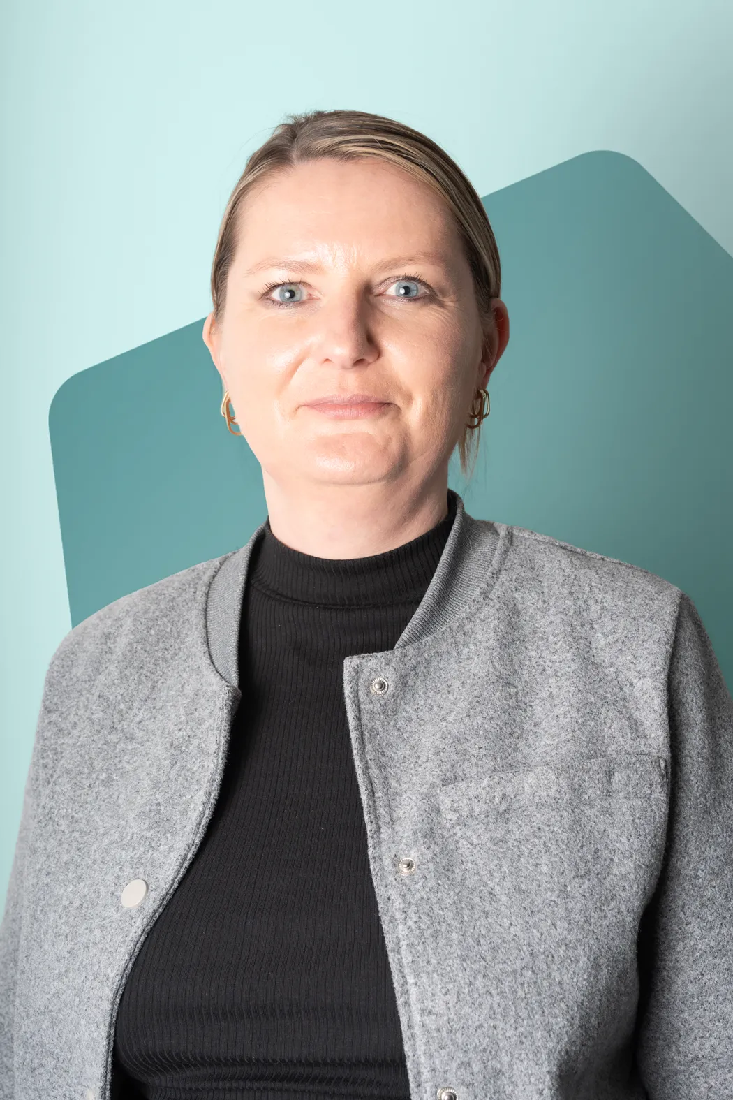 Portrait of a woman with blue eyes and blonde hair pulled back, wearing gold hoop earrings, a black ribbed top, and a gray buttoned jacket against a light blue background with a teal geometric shape.