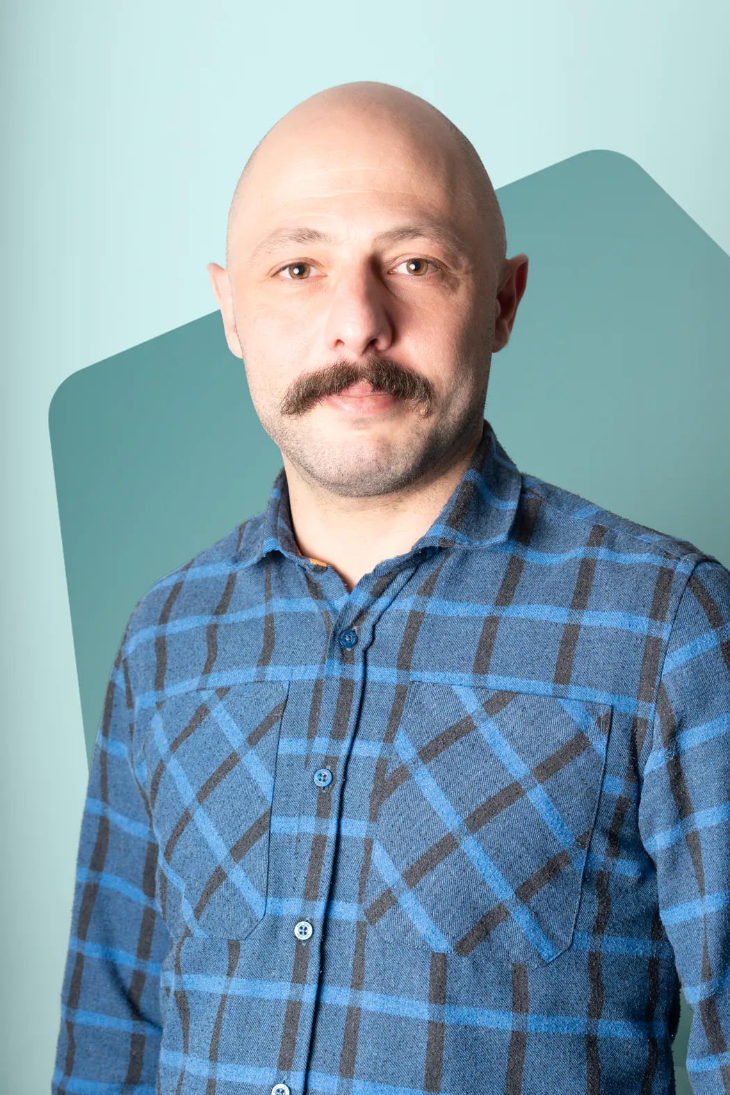 Portrait of a bald man with a mustache wearing a blue and black plaid shirt against a light blue background.