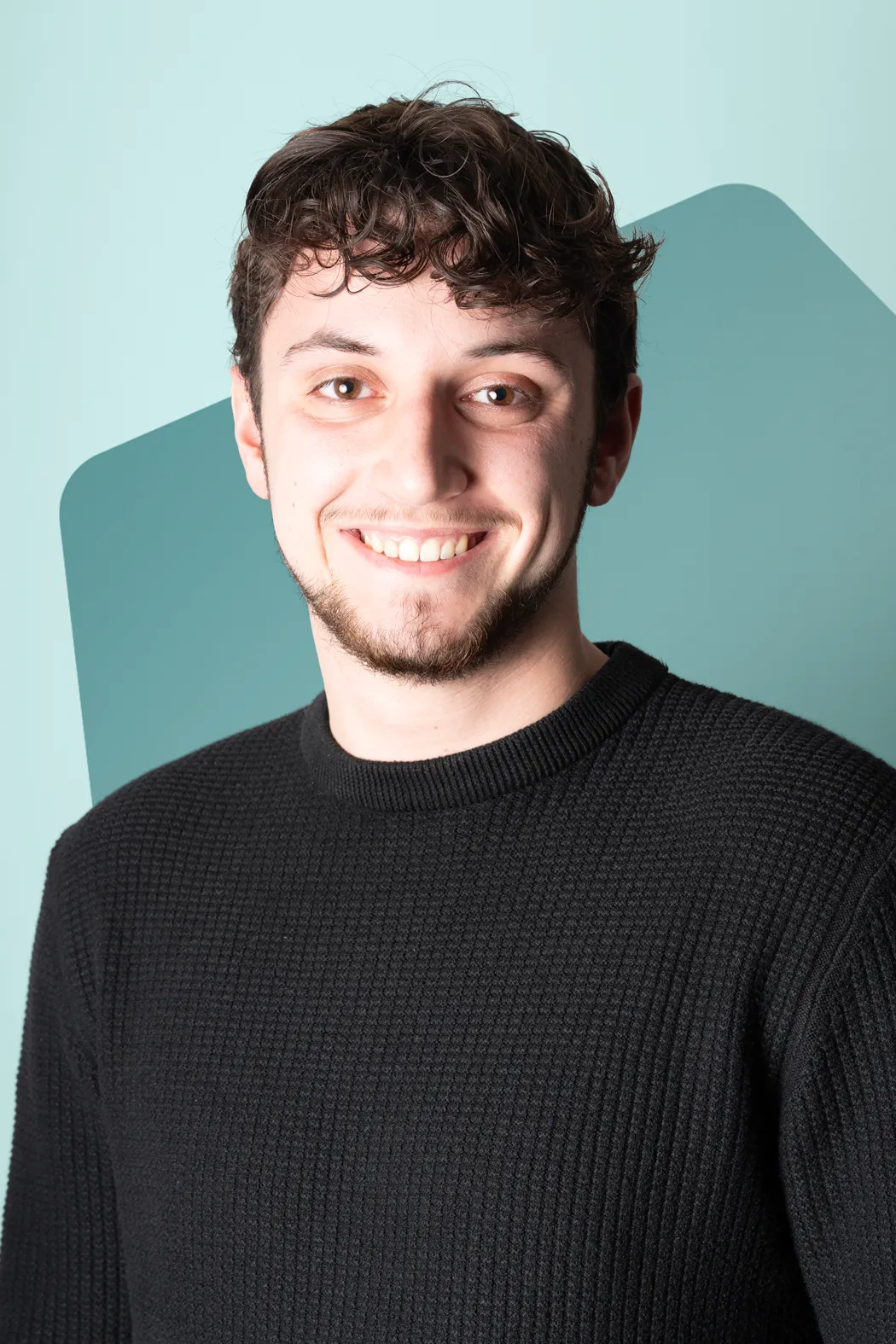 Smiling young man with short curly brown hair and light facial hair wearing a black sweater in front of a teal geometric background.