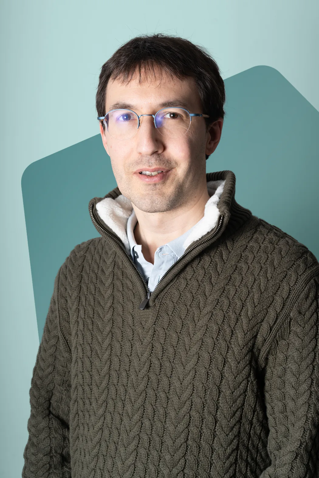 Man wearing transparent glasses and a thick olive green cable knit sweater with a fleece-lined collar.