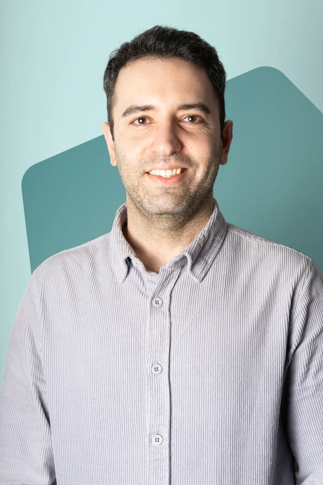 Smiling man with short dark hair wearing a light striped button-up shirt against a teal and light blue background.