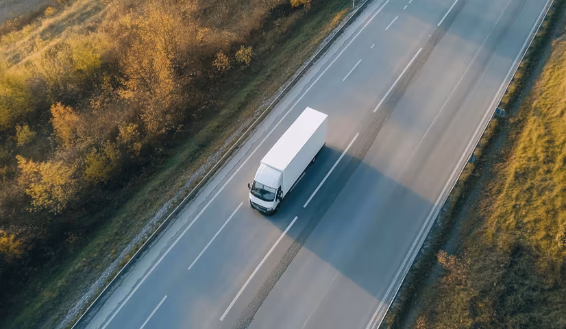 A white delivery truck driving on a two-lane highway surrounded by green and autumnal trees.