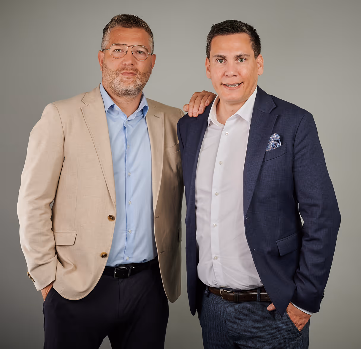 Zwei Männer in Anzügen stehen nebeneinander, einer mit heller Brille und beigefarbenem Sakko, der andere mit blauem Sakko und weißem Hemd, vor grauem Hintergrund.