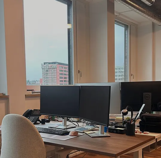 Moderner Schreibtisch mit zwei Monitoren, Tastatur, Maus, Telefon und Stuhl in der Nähe großer Fenster.