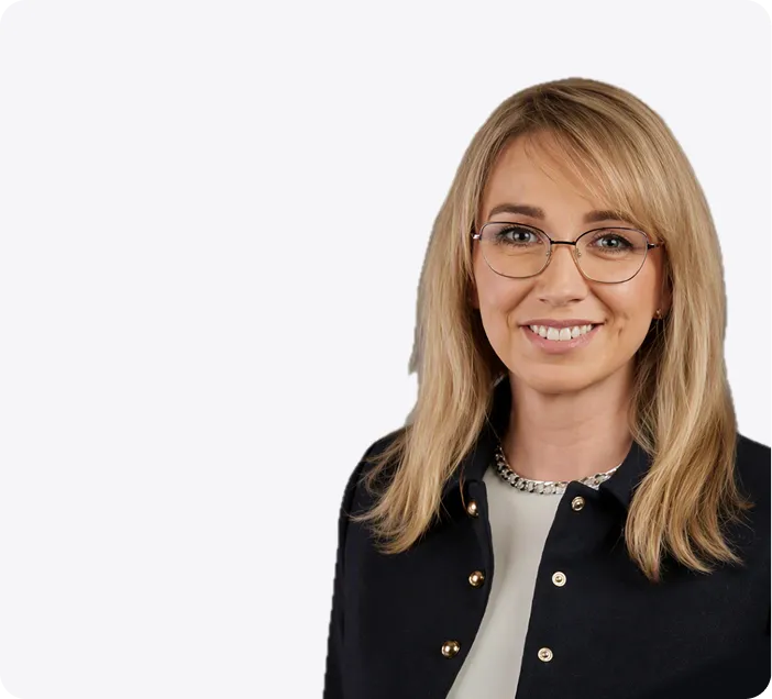 Ein Porträt einer lächelnder Frau mit blonden Haaren und einer Brille die eine schwarze Jacke und ein weißes Hemd angezogen hat