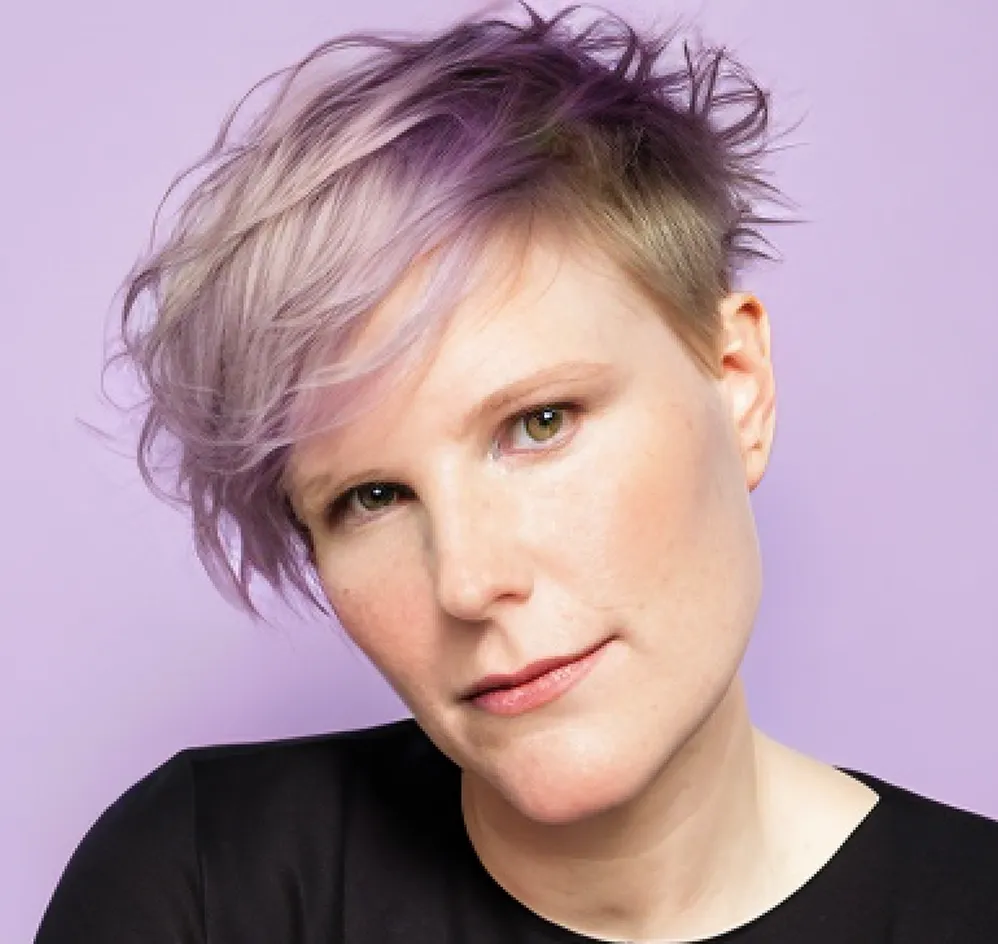 Das Portraitfoto zeigt eine weiße Person mit kurzen, hell-violett gefärbten und rot-blonden Haaren. Die Person ist weiß, hat den Kopf leicht nach links geneigt und guckt freundlich geradeaus in die Kamera. Die Person trägt ein schwarzes Shirt. Der Hintergrund ist hellviolett – ähnlich der Haarfarbe. Die abgebildete Person ist Daniela Schubert.