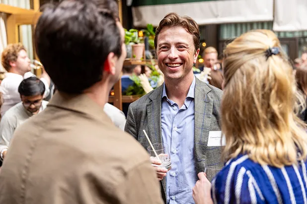 Smiling man in a gray blazer holding a drink and talking to two people at a social event.