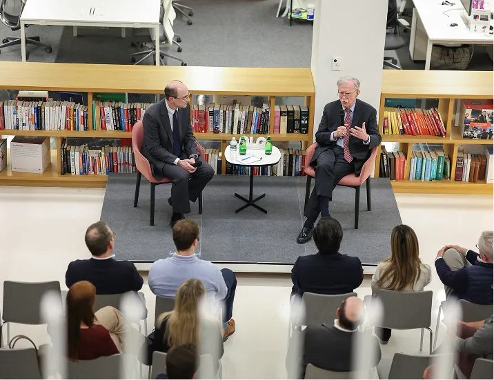 Two men in suits seated on stage having a discussion in front of an audience in a modern room with bookshelves.