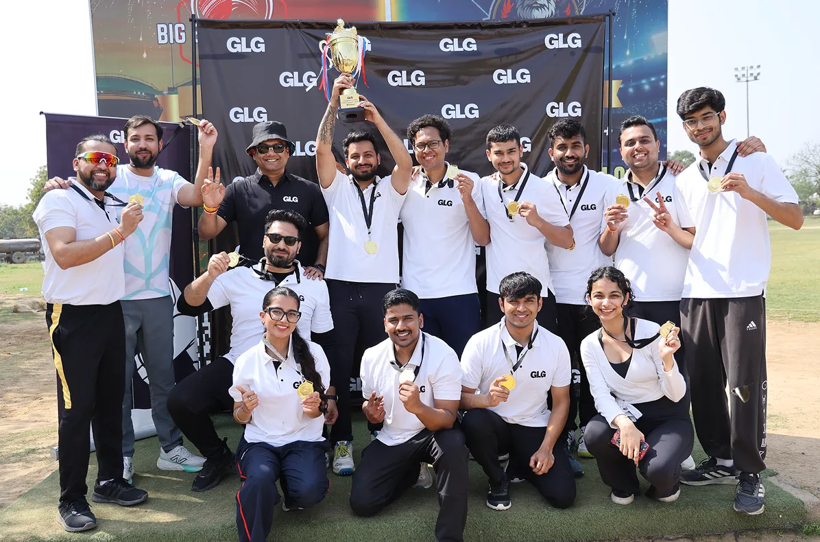 Group of 14 people wearing GLG shirts smiling and posing with gold medals and a trophy outdoors in front of a GLG backdrop.
