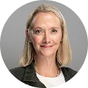 Portrait of a smiling woman with straight blonde hair wearing a black blazer and white top against a gray background.