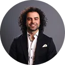 Smiling man with curly hair wearing a black blazer and white shirt against a grey background.