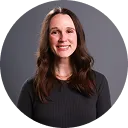 Smiling woman with long brown hair wearing a black top against a dark gray background.