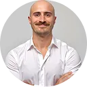 Smiling bald man with a mustache wearing a white button-up shirt, standing with arms crossed.