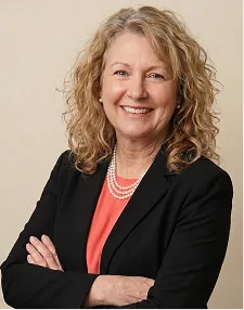 Smiling middle-aged woman with curly blonde hair wearing a black blazer, coral top, and pearl necklace, standing with arms crossed against a beige background.
