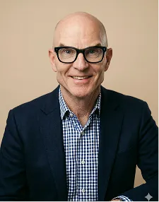 Smiling bald man wearing black glasses, a checked shirt, and a dark blazer against a beige background.