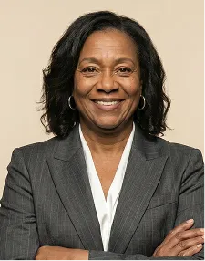 Confident middle-aged woman with curly hair wearing a gray blazer and white blouse, smiling with arms crossed.