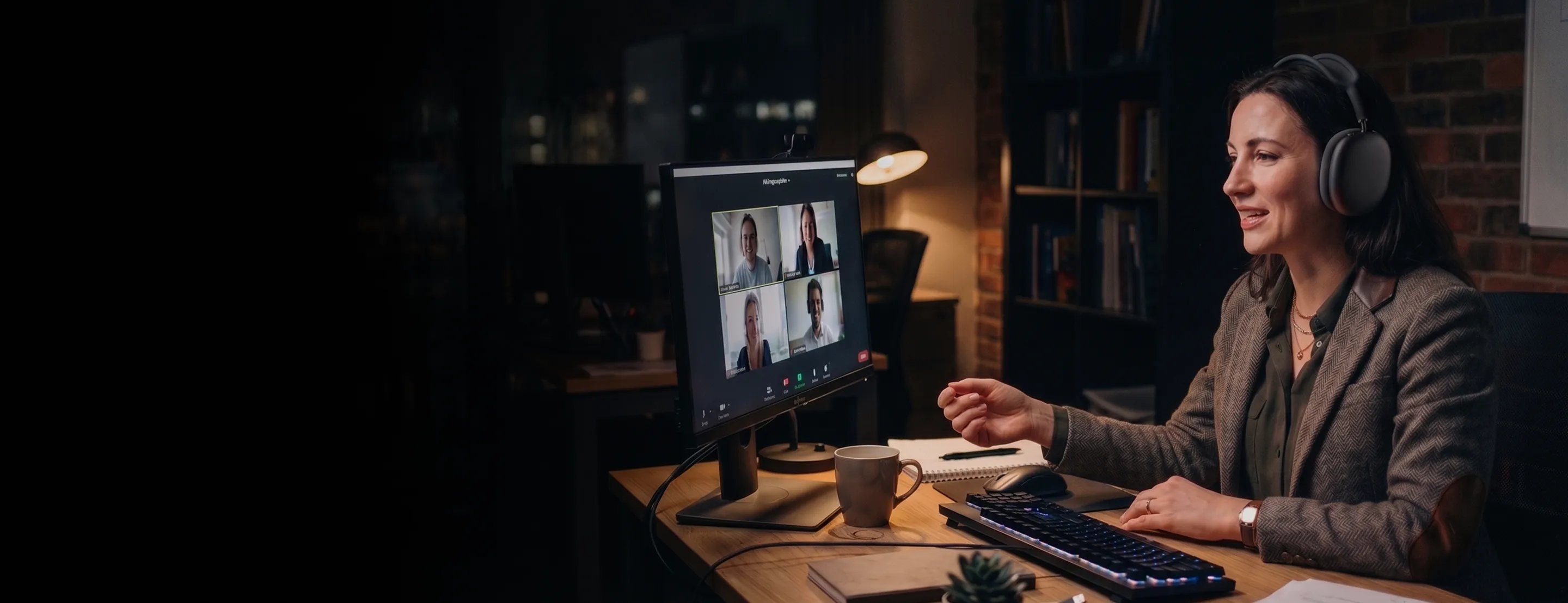 Woman wearing headphones sitting at a desk and participating in a video conference call on a computer monitor showing four people.