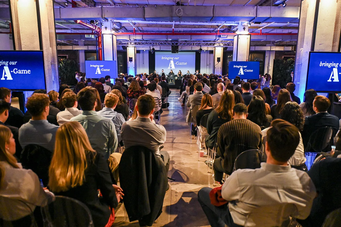 Audience seated in rows facing a panel discussion stage with screens displaying 'bringing our A-Game' in an industrial event space.