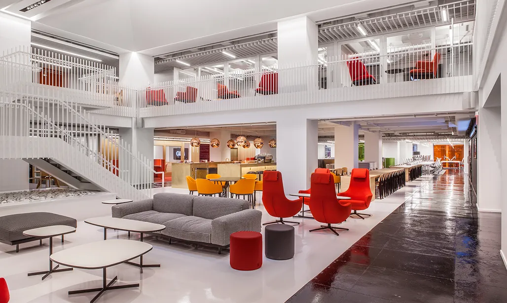 Modern office lounge with gray sofa, red and orange chairs, white tables, and a staircase leading to a mezzanine with red seating.