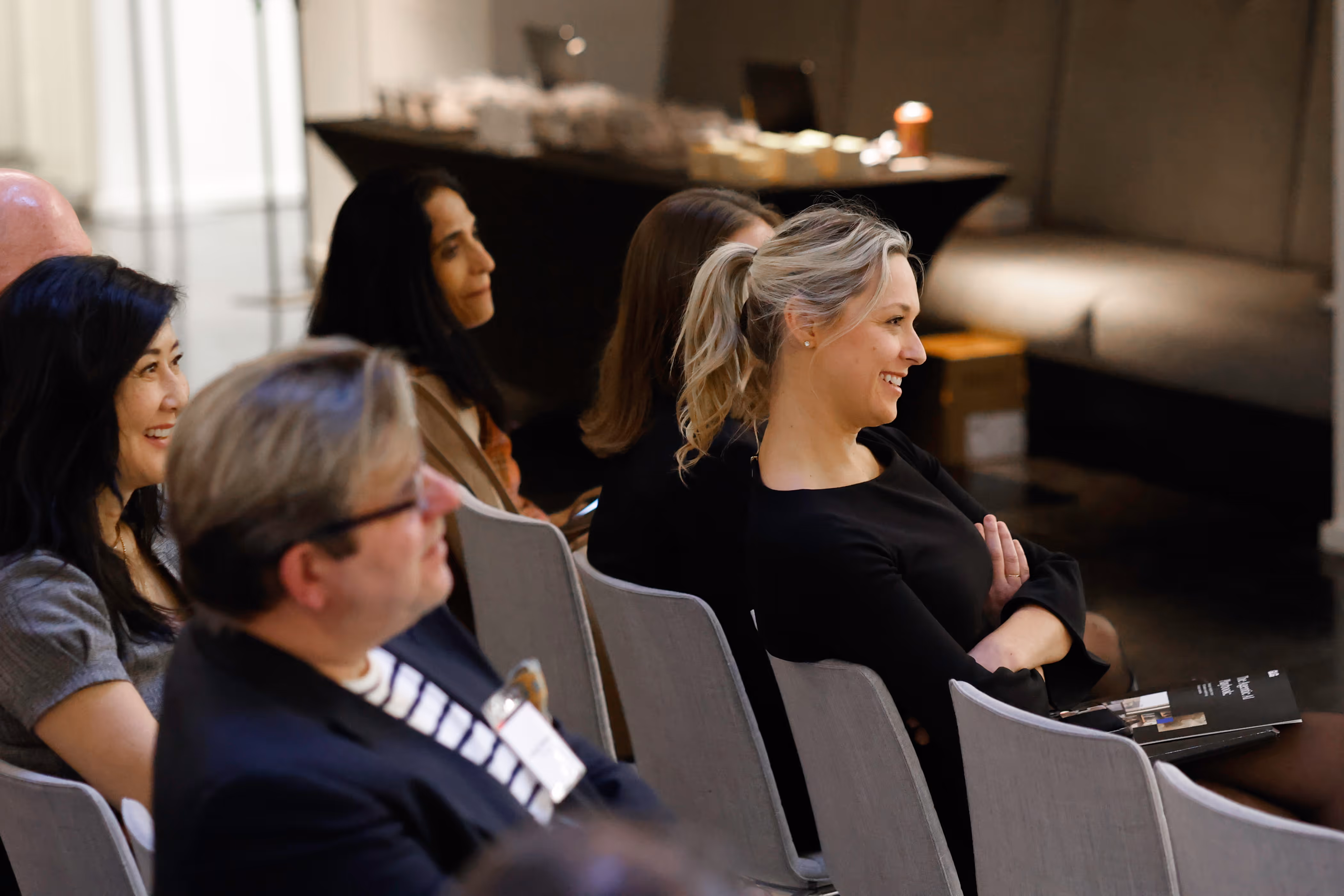 People seated in rows attentively watching an event, with a smiling blonde woman in the foreground.